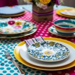 A table set with various patterned fabrics and objects, such as floral plates, striped napkins, and polka dot tablecloth